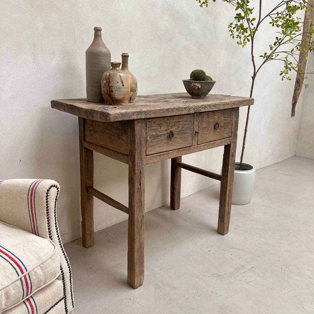 A rustic wooden console table with two drawers and a weathered finish, styled with ceramic vases and a small bowl on top, set against a light textured wall with a potted plant nearby.