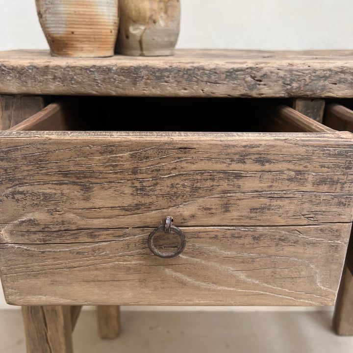 Close-up of a rustic wooden drawer with a distressed finish and a dark metal ring pull, with part of a matching wooden surface and two ceramic vases visible above.