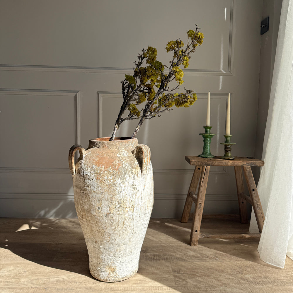 Large textured vase with dried flowers on a wooden floor, with a small wooden stool and candles in the background.