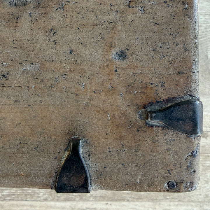 Close-up of the corner of a rustic wooden board or box with dark metal corner protectors, showing a weathered brown wood grain surface.
