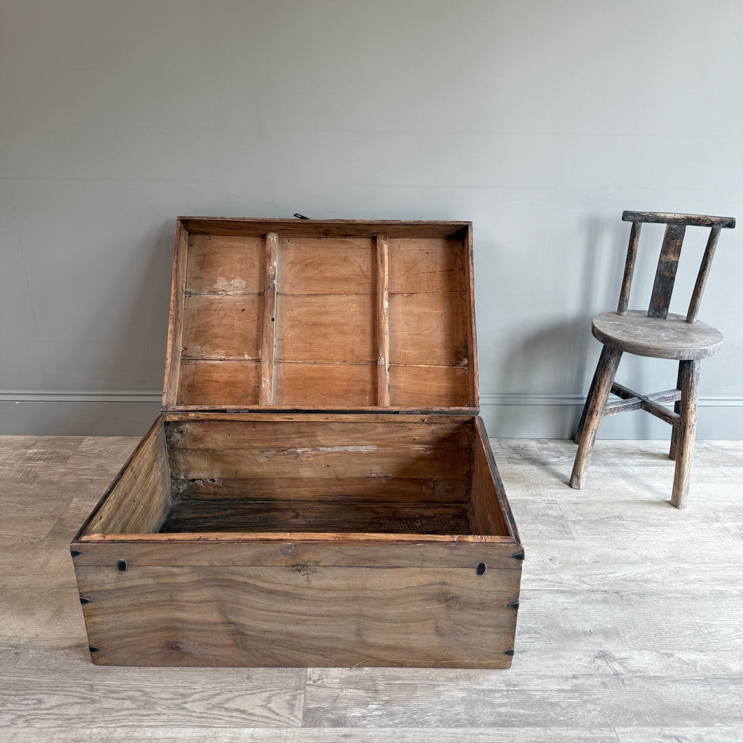 A large rustic wooden storage chest with its lid open, set on a light wood floor against a gray wall, with a small wooden chair to the right.