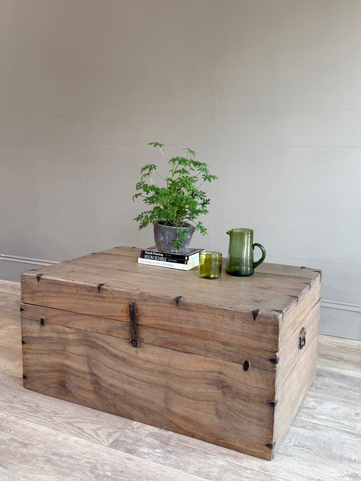 A rustic wooden storage chest with metal corner brackets and a latch, styled on a light wood floor against a neutral beige wall with a small potted plant and glassware on top.