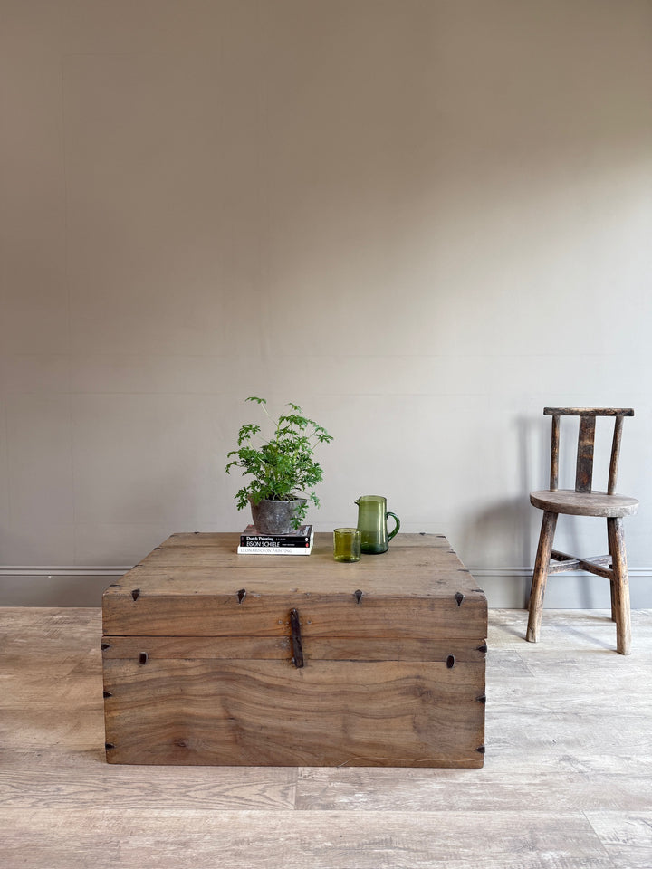 A rustic wooden storage chest with metal corner brackets and a latch, styled with a small potted plant and glasses on top, set in a room with a beige wall and light wood floor next to a wooden chair.