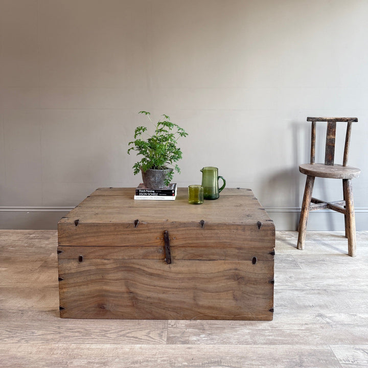 A rustic wooden storage chest with metal corner brackets and a latch, styled with a small potted plant and glasses on top, set in a room with a beige wall and light wood floor next to a wooden chair.