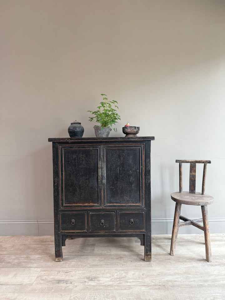 A dark distressed wooden cabinet with two doors and three drawers, styled with small decorative pots and a plant on top, next to a rustic wooden chair against a plain beige wall.