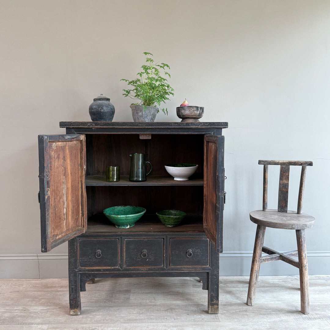 A rustic dark wood cabinet with its doors open, styled with ceramic bowls, a green plant, and a small bowl on top, set next to a matching wooden chair against a neutral wall.