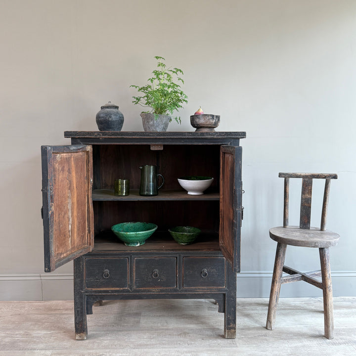 A rustic dark wood cabinet with its doors open, styled with ceramic bowls, a green plant, and a small bowl on top, set next to a matching wooden chair against a neutral wall.