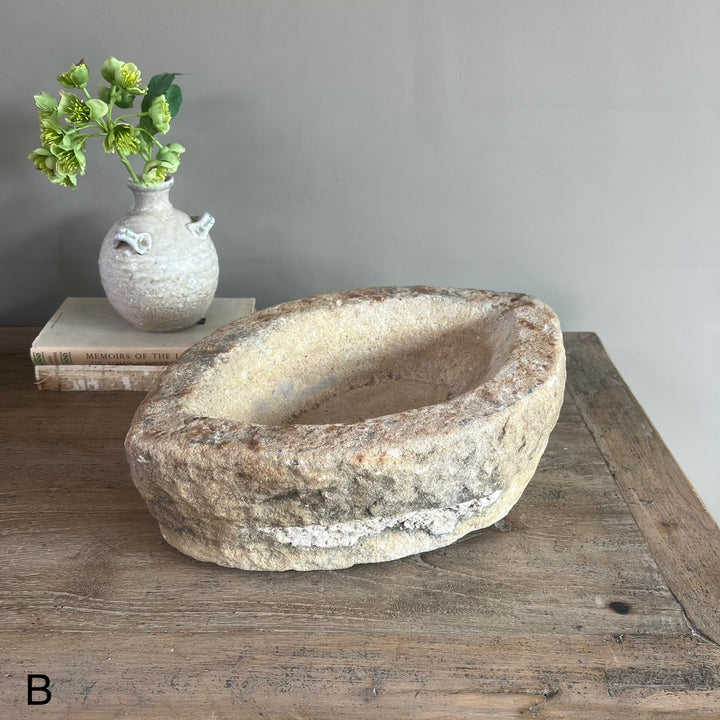 Decorative stone bowl on a wooden surface with a vase of greenery in the background.