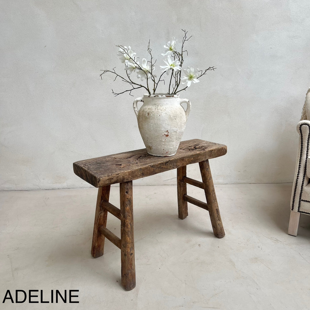 Vintage wooden stool with a ceramic vase and white flowers against a plain wall.