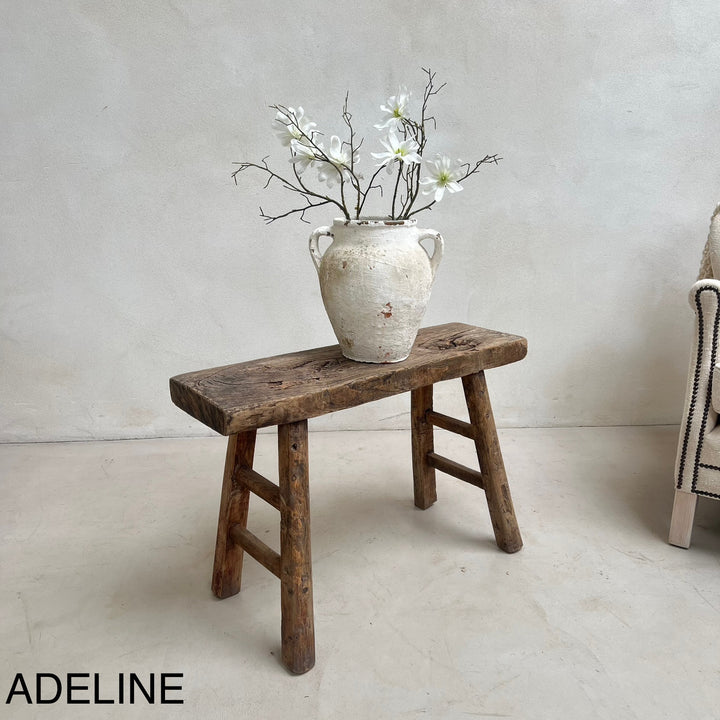 Vintage wooden stool with a ceramic vase and white flowers against a plain wall.