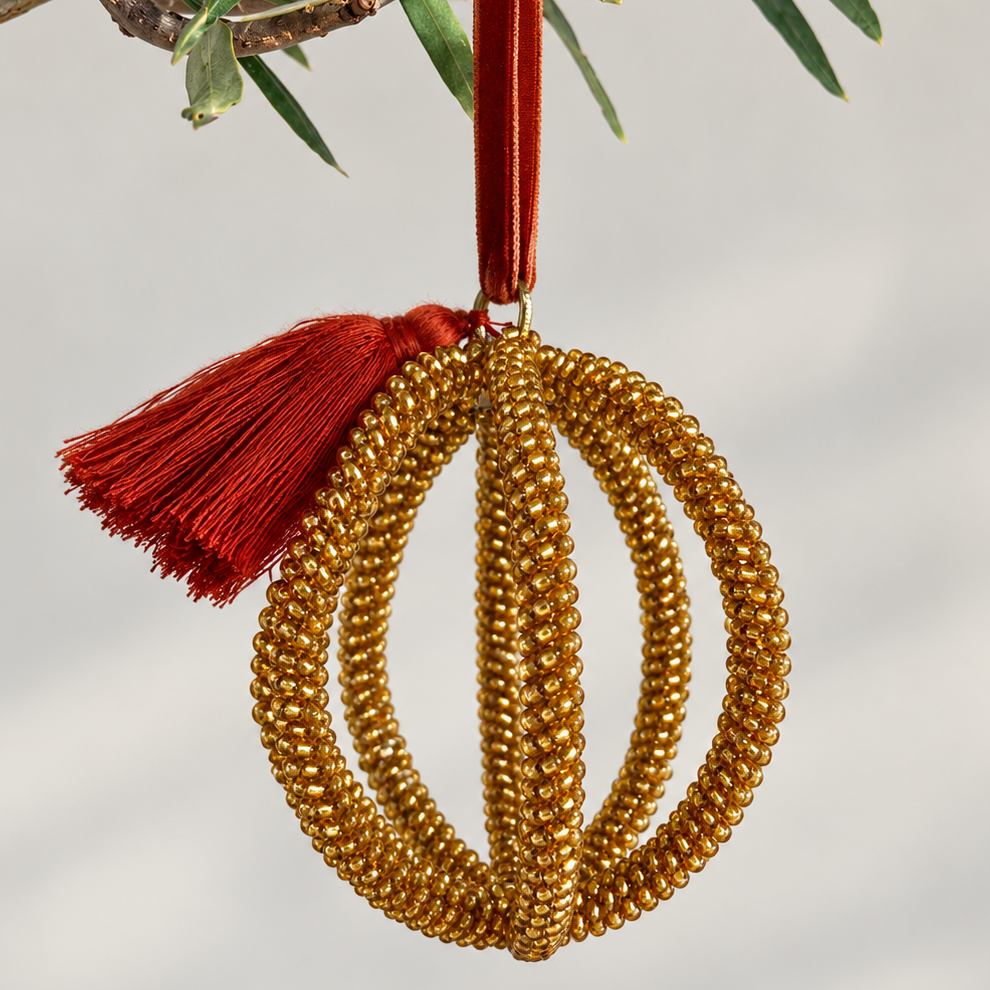 A gold beaded spherical Christmas ornament with a red tassel and hanging ribbon, shown hanging from a branch against a soft gray background.