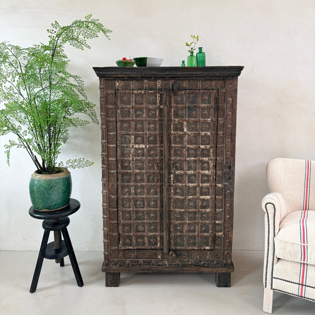 indian antique cabinet with decorative items against a white wall, with a plant and chair in the foreground.