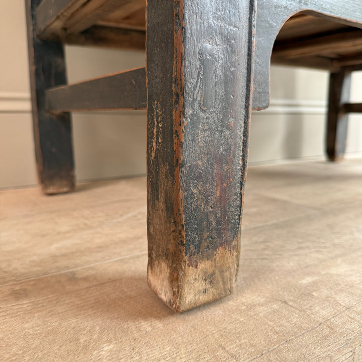 Close-up of a weathered dark wood furniture leg with chipped black paint and exposed brown wood, standing on a light wood floor.