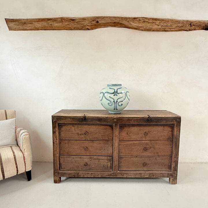 Wooden dresser with a vase on top in a room with a beige armchair and decorative wooden beam.