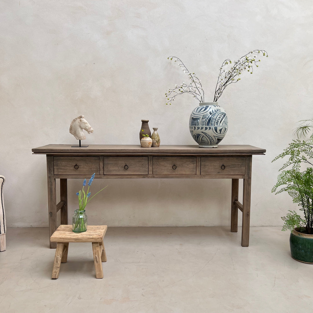 A rustic gray-brown wooden console table with three drawers and round knobs, styled with decorative vases and a small wooden stool in a minimalist room with a textured plaster wall.