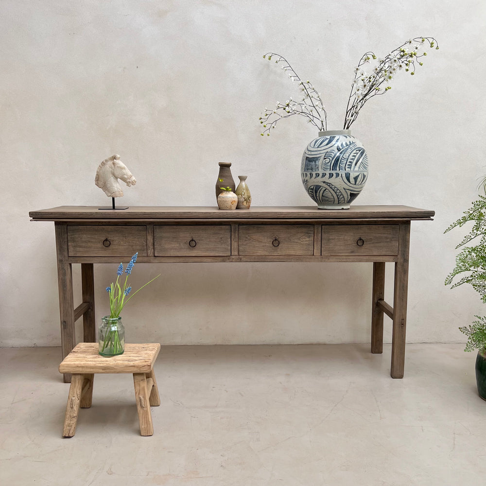 A rustic gray wooden console table with five drawers, styled with decorative vases, a small wooden stool, and a horse head sculpture against a textured plaster wall.