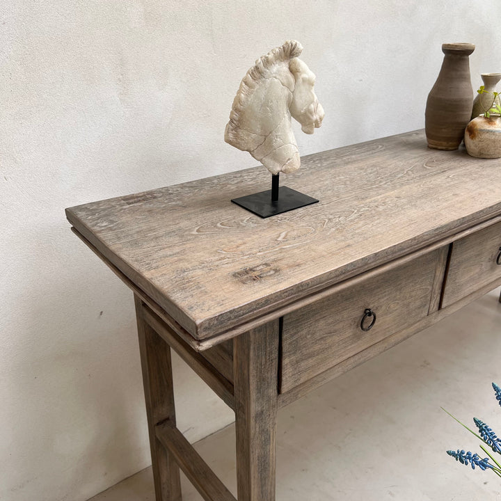 A rustic gray-brown wooden console table with drawers and ring pulls, styled with a horse head sculpture on a black stand and ceramic vases against a textured off-white wall.
