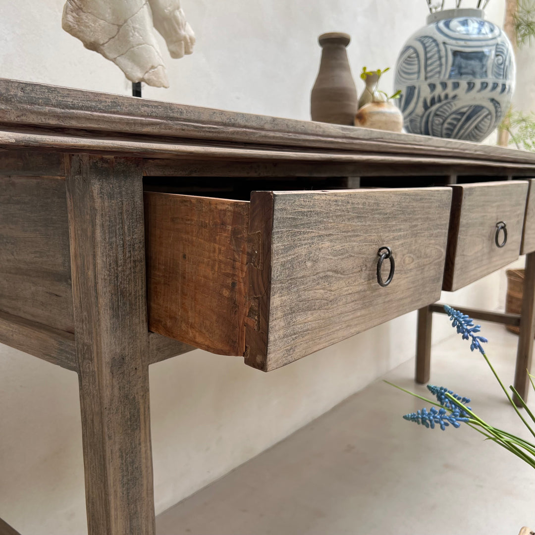 A rustic wooden console table with weathered gray-brown finish and dark metal ring pulls on its drawers, shown in a styled interior setting with decorative vases and flowers.