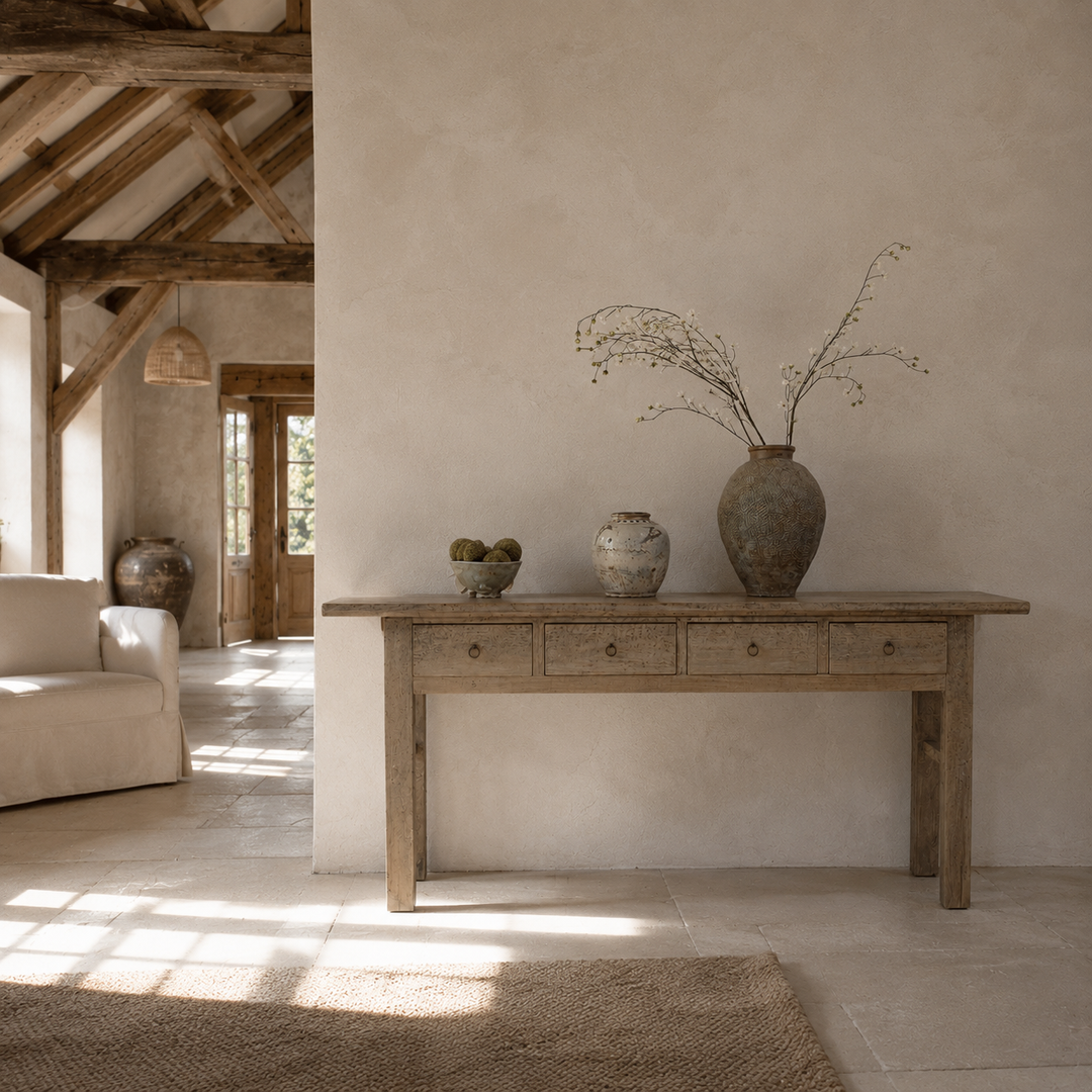 A rustic wooden console table with five drawers stands against a textured beige plaster wall in a bright, sunlit room. On top of the table are three ceramic vases, including a large vase with dried branches, alongside a light-colored armchair and a rug in the foreground.