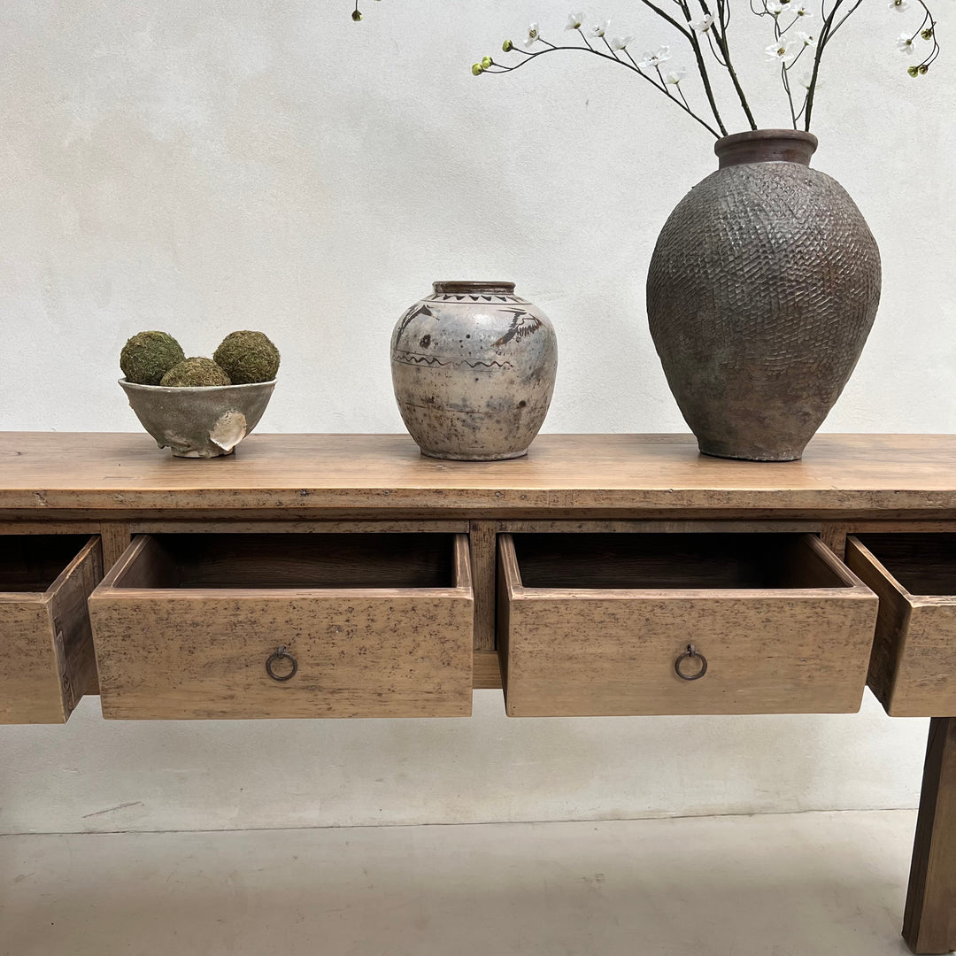 A rustic light brown wooden console table with three open drawers, styled with a textured gray ceramic vase, a small bowl of mossy decorative spheres, and a larger dark textured vase with dried branches against a neutral off-white wall.
