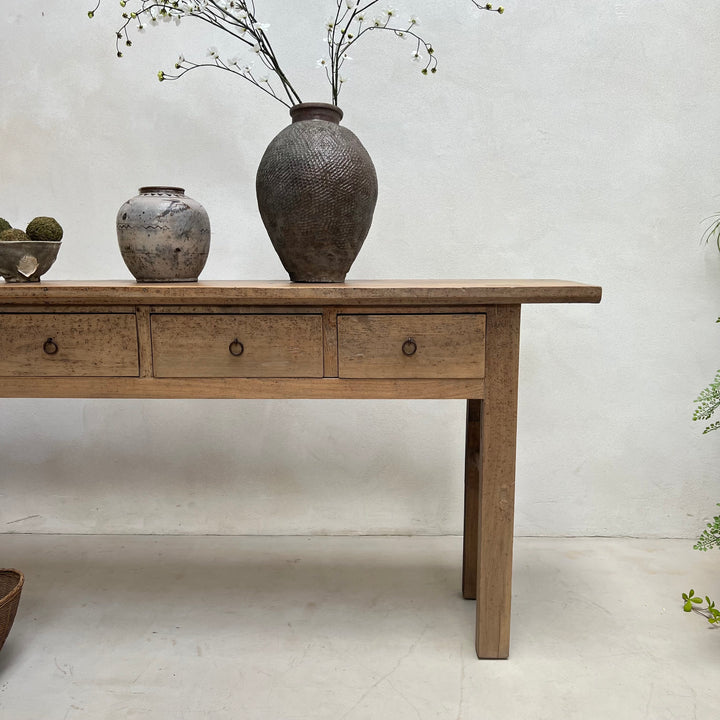 A rustic light brown wooden console table with three drawers and round metal knobs, styled with ceramic vases on top against a textured off-white wall.