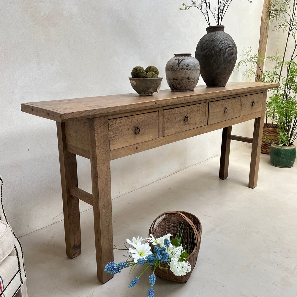 A rustic light brown wooden console table with five drawers and small round metal knobs, styled with ceramic vases and a basket of flowers in a neutral indoor setting.