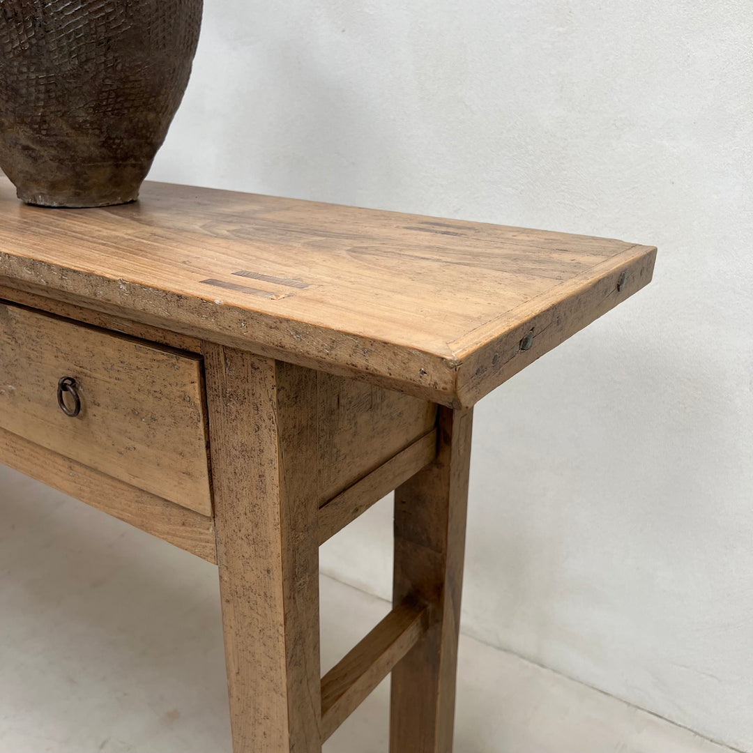 A rustic light brown wooden console table with a single drawer and a dark woven ceramic vase on top, set against a plain white wall.