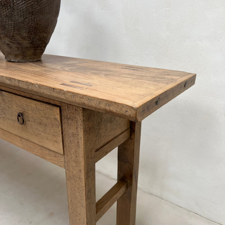A rustic light brown wooden console table with a single drawer and a dark woven ceramic vase on top, set against a plain white wall.