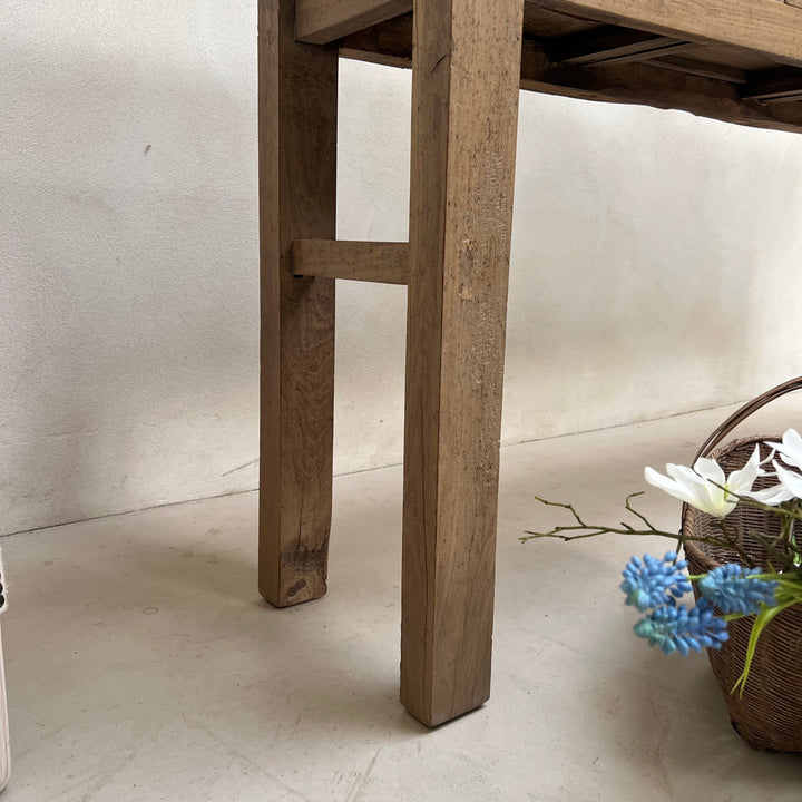 Close-up of a rustic wooden table leg with a crossbrace, set against a neutral off-white wall and floor, with a wicker basket and blue flowers in the lower right corner.