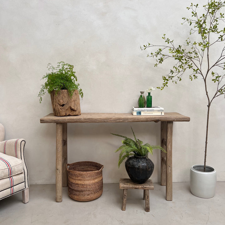 A rustic wooden console table styled in a minimalist room setting, featuring a potted green plant in a carved wood pot, books, and a small stool with another plant. The background is a textured light gray wall.