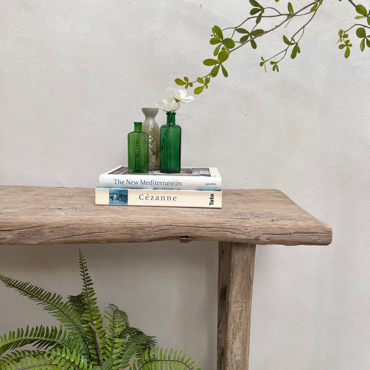 A rustic wooden console table styled with two green glass bottles and a small ceramic vase stacked on books, with a white flower, against a textured light gray wall with green foliage in the foreground and background.