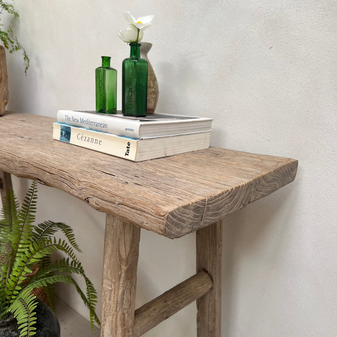 A rustic wooden console table with a thick rectangular top and thick legs, styled with two green glass bottles, a small vase with a white flower, and a stack of books against a light textured wall.