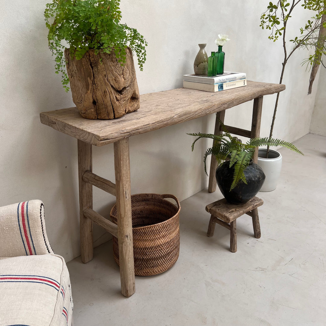 A rustic wooden console table with a weathered light brown finish, styled in a minimalist room with potted ferns, a woven basket, and a small stool.