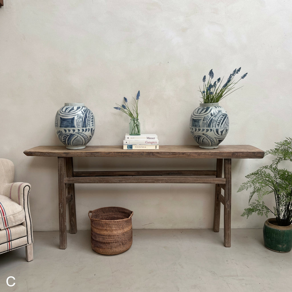 A rustic wooden console table styled in a minimalist room setting, featuring two blue and white patterned ceramic vases, a small stack of books, and a vase of lavender. A woven basket sits on the floor beneath the table.