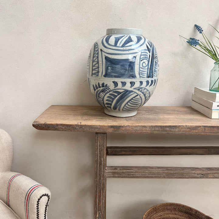 A white ceramic vase with a blue hand-painted abstract pattern sits on a rustic wooden console table against a textured beige wall.