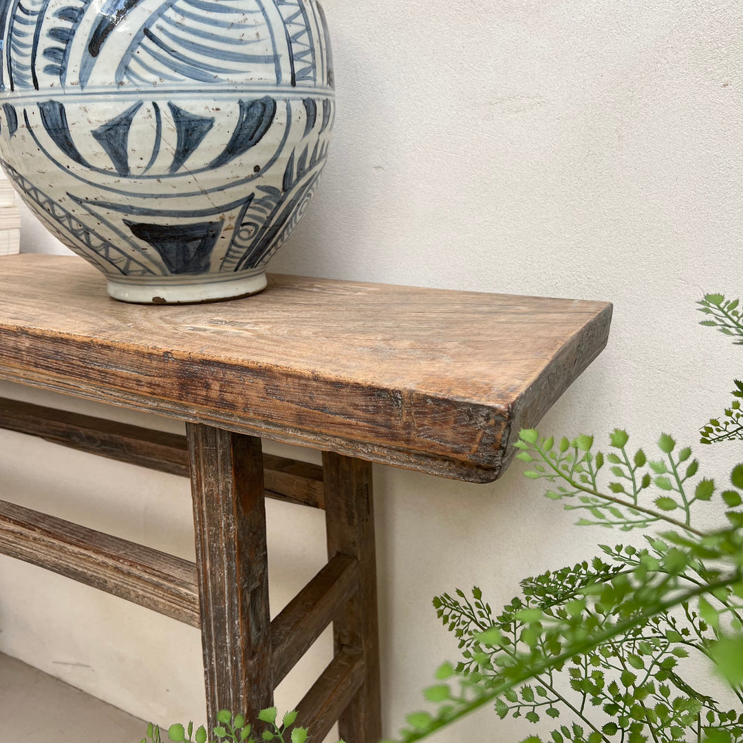 A rustic dark brown wooden console table with a thick top and slatted legs, styled with a blue and white patterned ceramic vase on top against a textured off-white wall, with green foliage in the foreground.