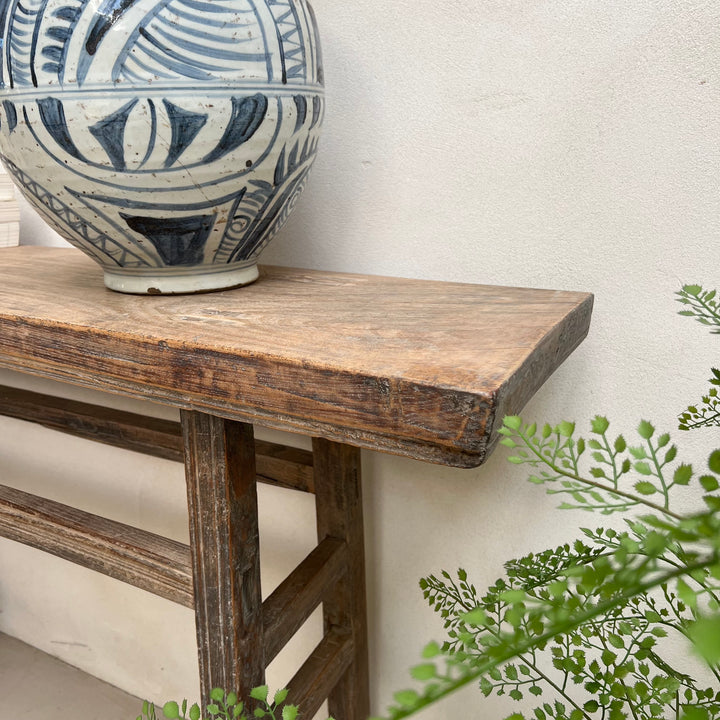 A rustic dark brown wooden console table with a thick top and slatted legs, styled with a blue and white patterned ceramic vase on top against a textured off-white wall, with green foliage in the foreground.