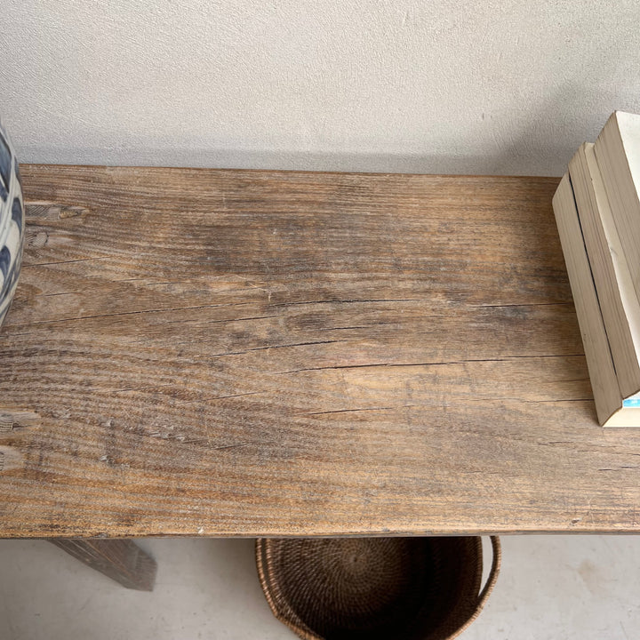 A rustic brown wooden tabletop or shelf with visible wood grain, shown in a real interior setting against a light wall. Part of a patterned vase appears on the left, and a stack of books and a woven basket are visible on the right and underneath.