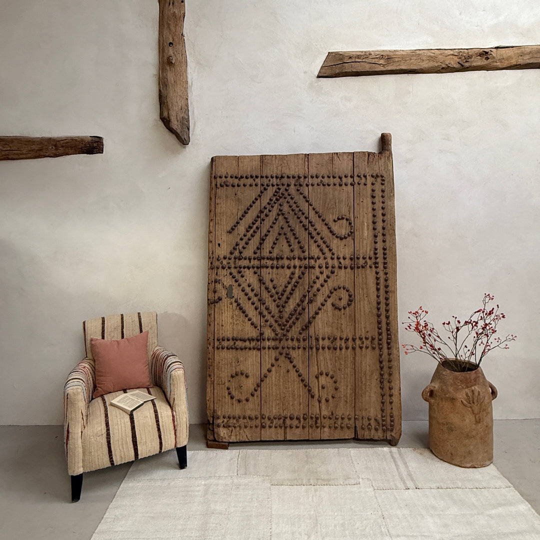 Decorative rug with geometric pattern on a white wall, with a chair and vase in the foreground.