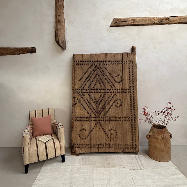 Decorative rug with geometric pattern on a white wall, with a chair and vase in the foreground.