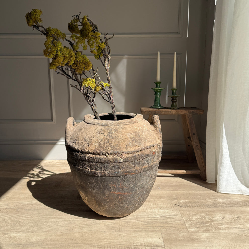 Large rustic pot with plants on a wooden floor in a room with candles and a curtain.