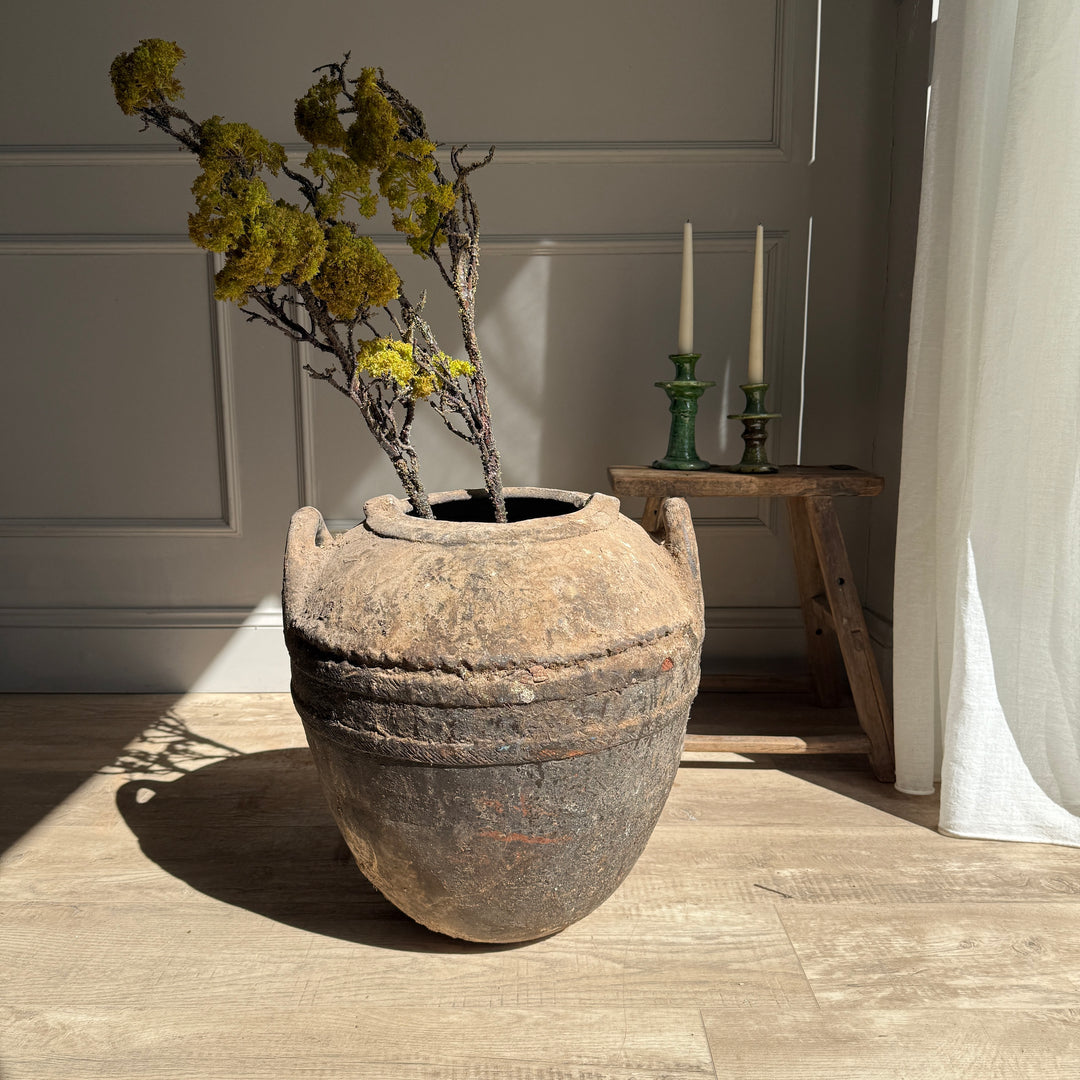 Large rustic pot with plants on a wooden floor in a room with candles and a curtain.