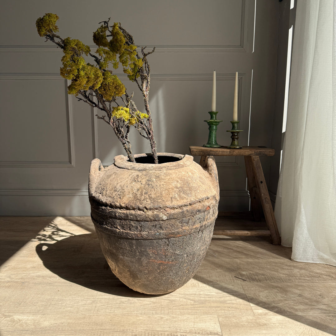 Vintage-style pot with dried plants on a wooden floor, with a small table and candles in the background.