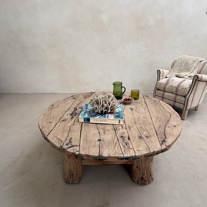 Round wooden table with decorative items in a room with a chair and light-colored floor.