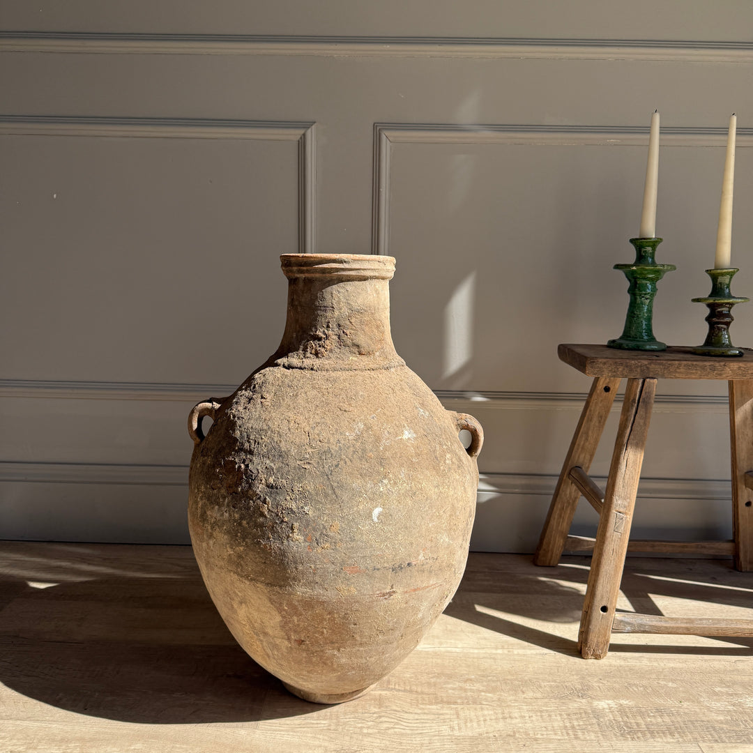 Large rustic vase on a wooden floor with a neutral wall and small wooden table in the background.