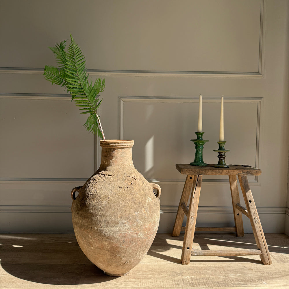 Large rustic vase with a plant next to a wooden stool with candles against a neutral wall.