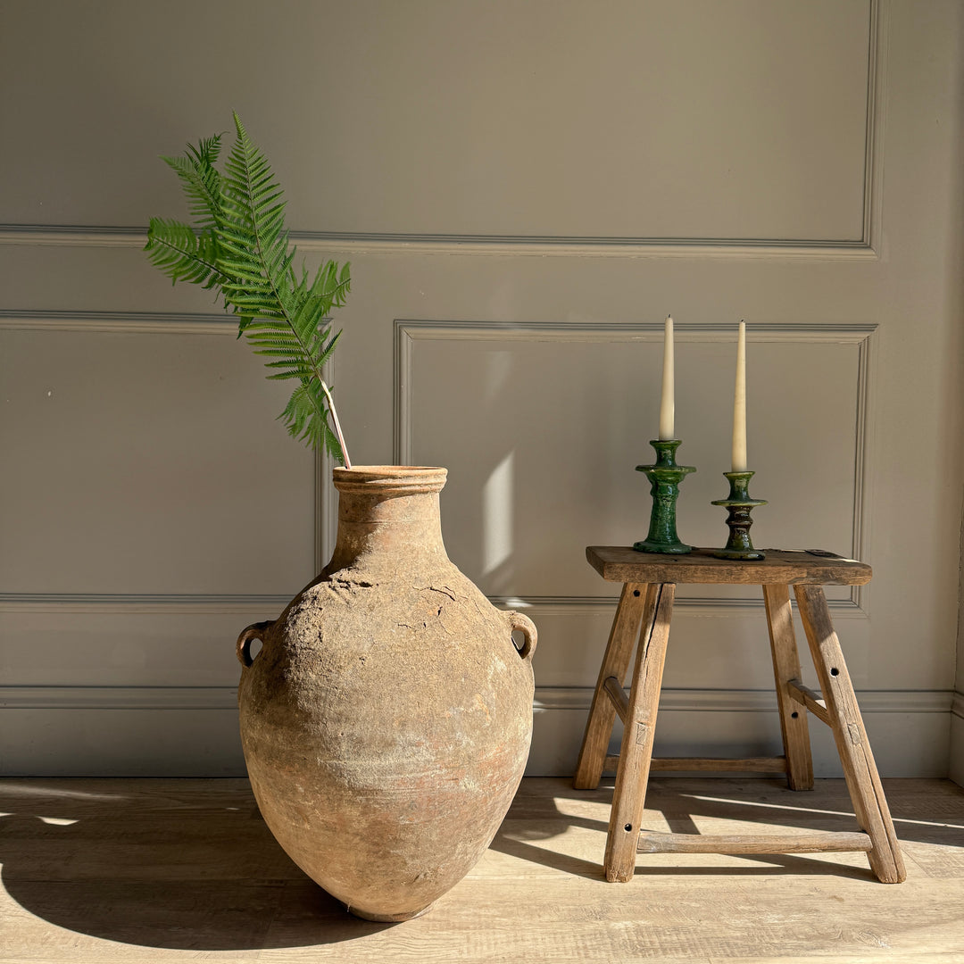 Large rustic vase with a plant next to a wooden stool with candles against a neutral wall.