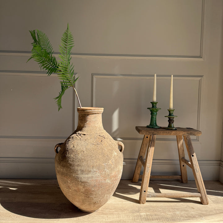 Large ceramic vase with a plant next to a wooden stool with candles against a neutral wall.