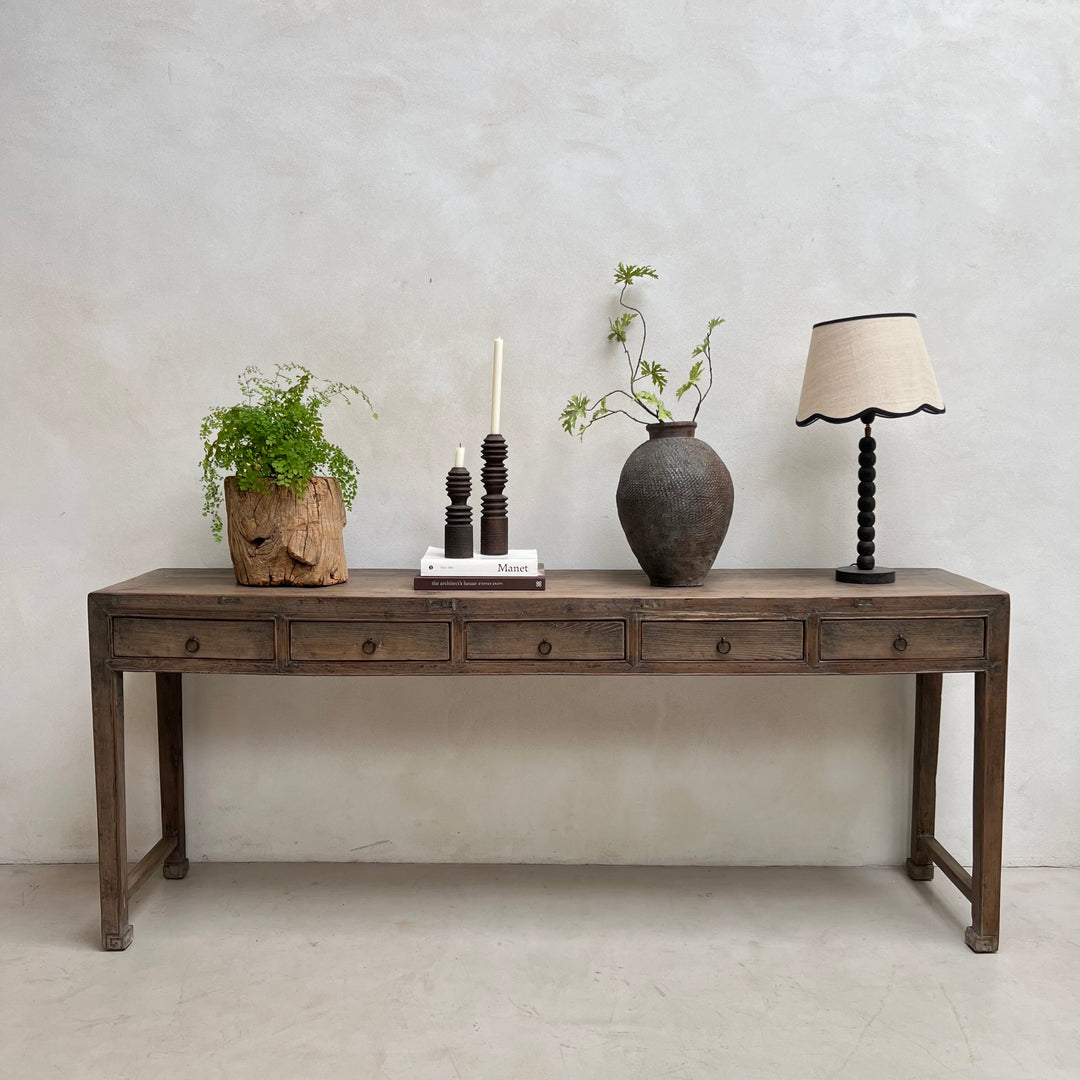 A rustic dark brown wooden console table with five drawers, styled with a potted green plant, a dark ceramic vase with branches, a candle holder, and a table lamp against a textured light gray wall.