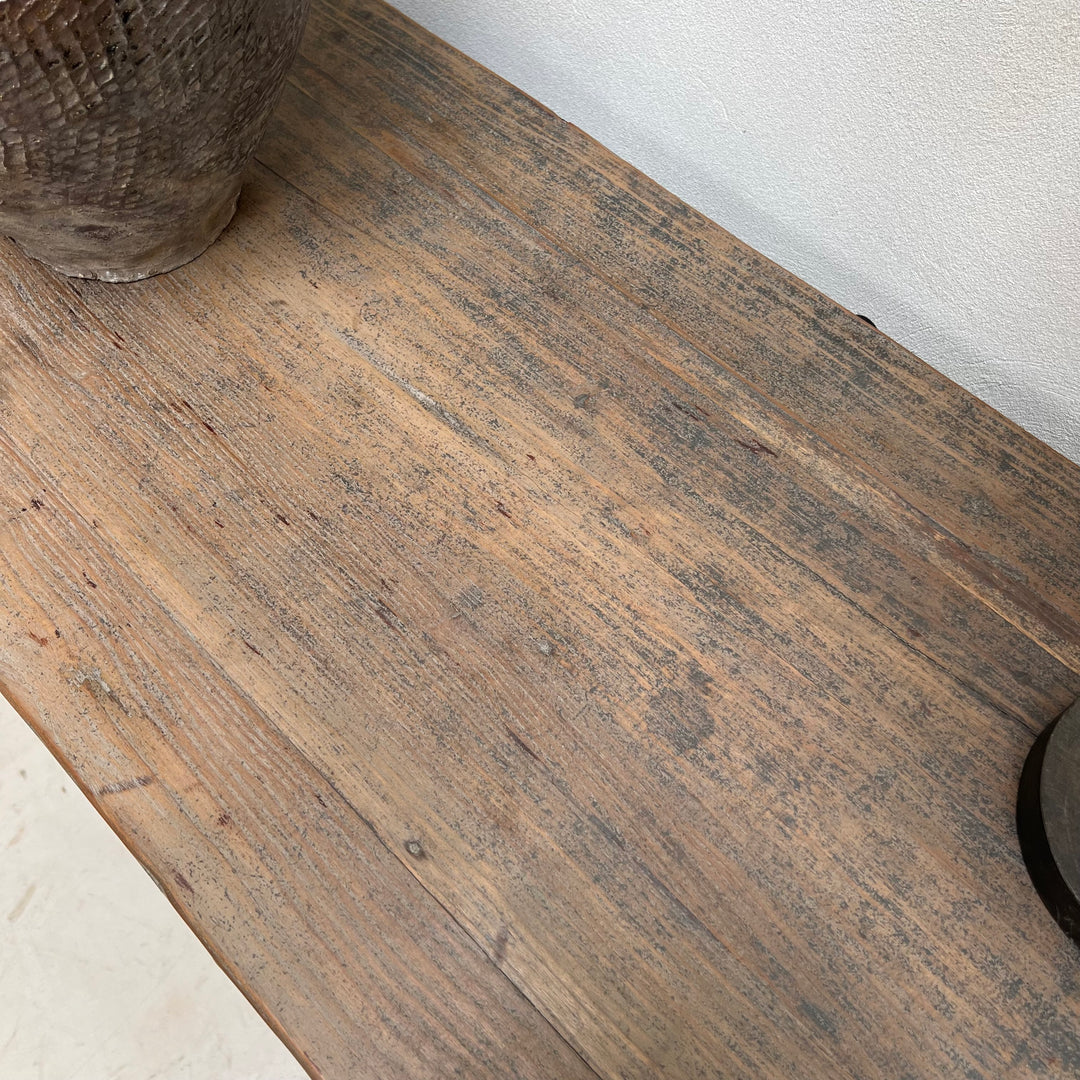A close-up of a rustic brown wooden tabletop with visible grain and weathered texture, with parts of two decorative objects visible at the corners against a light wall.